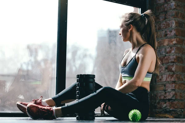 En flicka i sportkläder sitter i studion på golvet efter ett spänt träningspass, bredvid henne finns en massage cylinder och myofascial boll. — Stockfoto