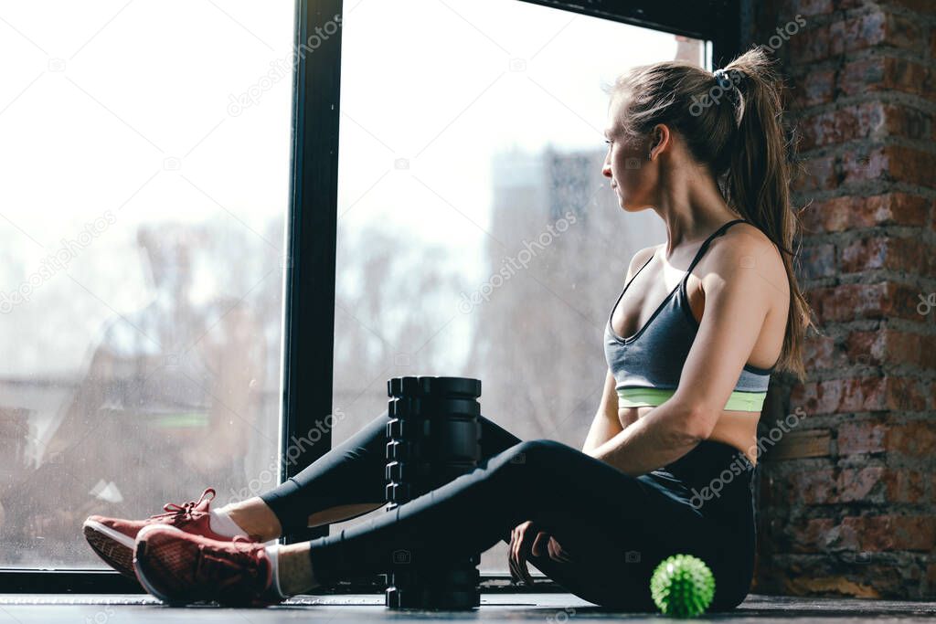 A girl in sportswear is sitting in the studio on the floor after a tense workout, next to her are a massage cylinder and myofascial ball.