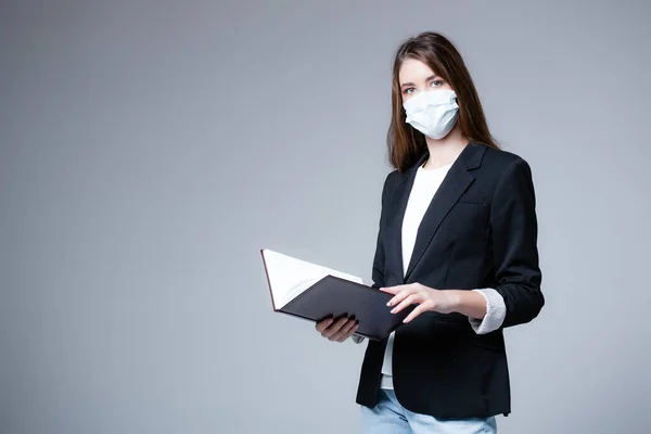 Menina em uma máscara médica e uma jaqueta preta com belos olhos segurando datebook no fundo branco — Fotografia de Stock