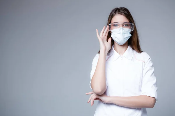 Médico estudante russo está em um casaco branco em uma máscara médica e óculos em um fundo branco durante uma pandemia — Fotografia de Stock