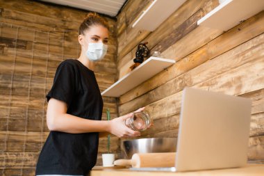 Mutfakta maskeli bir kız internette yemek yapmayı öğreniyor ve dizüstü bilgisayarına bakıyor. Karantina modunda aşçılık eğitimi