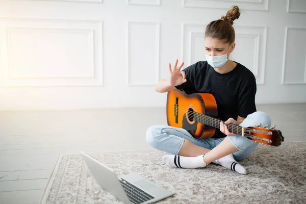 A quarantined masked girl practices guitar lessons on a laptop through a video conference with a teacher in a bright room.