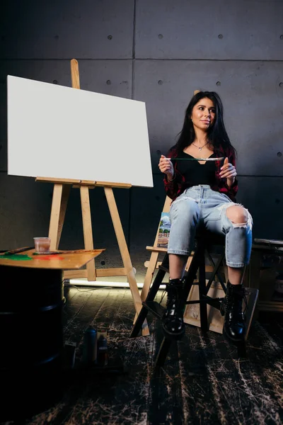 Mock Up Canvas en blanco. Morena artista en una camisa a cuadros rojo y jeans, acaba de terminar de pintar una nueva pintura acrílica abstracta . — Foto de Stock