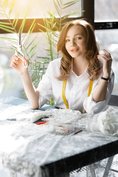 Sweetheart seamstress sitting behind a Desk and makes custom wedding lace dress and holding a pair of scissors on the background of sunlight