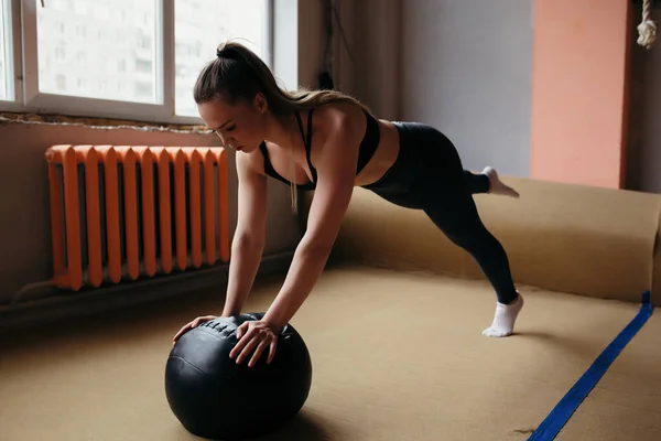 A menina instrutora de fitness fazendo Pilates na bola de medicina no ginásio. Vestido com um top preto e leggings — Fotografia de Stock