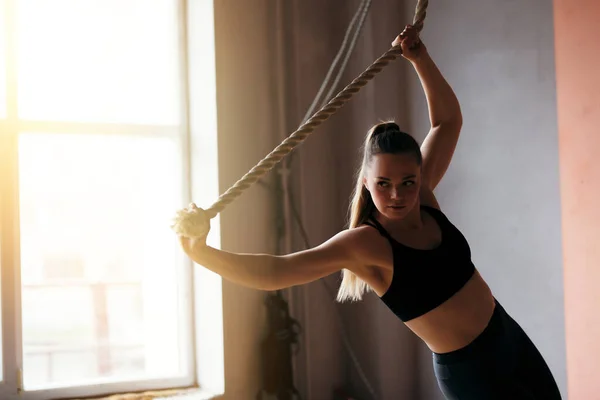 Sportig flicka gör övningar med en sport hängande rep i en solig fitness Studio nära fönstret. Klädd i svart topp och leggings — Stockfoto