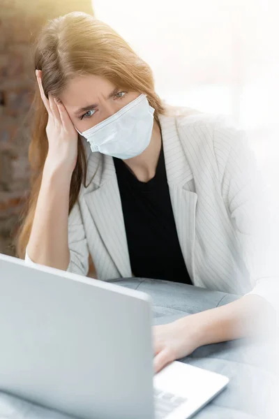 Mujer en una chaqueta y máscara médica se ve perplejo en la pantalla de las computadoras portátiles tratando de resolver un problema de trabajo. Trabajo en casa . — Foto de Stock