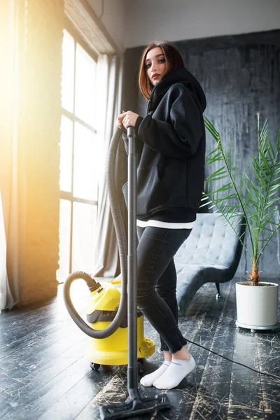 A woman in a black hoodie is vacuuming the house and dancing while cleaning. Yellow vacuum cleaner, panoramic windows, loft studio