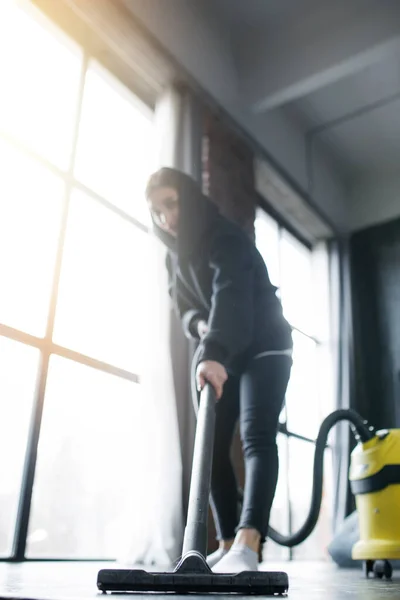 Stylish housewife in a black hoodie vacuums and thoroughly removes dirt. Yellow vacuum cleaner, panoramic windows, loft studio. — Stock Photo, Image