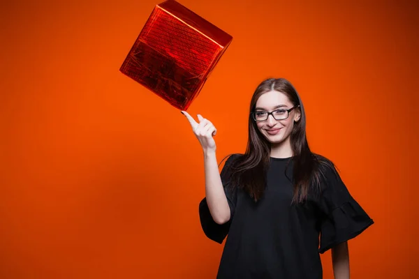 Mulher de vestido preto segura uma caixa vermelha com um dedo e sorri para a câmera. Vendas e descontos com fundo laranja . — Fotografia de Stock