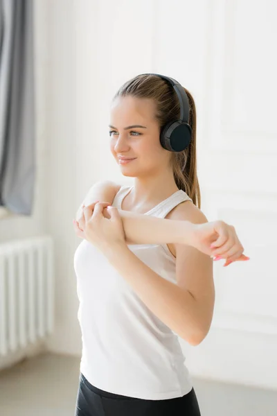 A girl of European appearance smiles, stands in black headphones and does shoulder stretching exercises in a bright studio — Stock Photo, Image