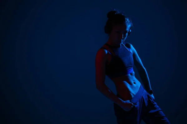 Uma menina com músculos abdominais em relevo iluminados por poses de néon azul contra um fundo escuro e olha para a câmera . — Fotografia de Stock