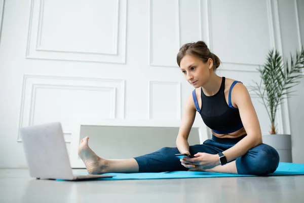 O femeie se uită acasă la o lecție de fitness pe un laptop și ține un smartphone în mână . — Fotografie, imagine de stoc