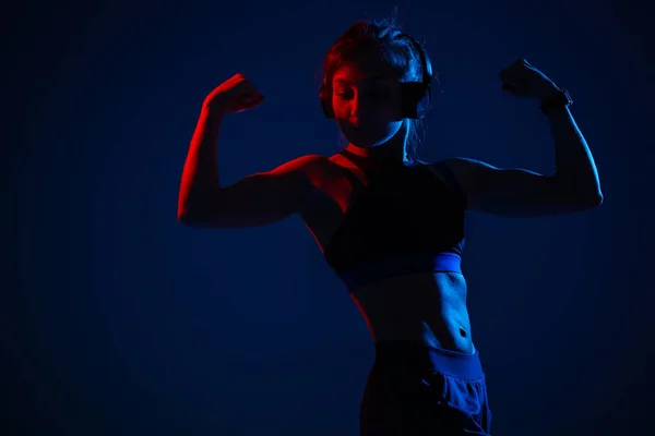 Uma menina com alívio iluminado músculos abdominais mostra bíceps. Desportista em fones de ouvido em uma luz vermelha e azul néon . — Fotografia de Stock