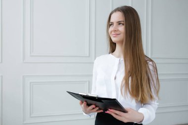 Beyaz tişörtlü Avrupalı bir kız elinde kağıttan bir tablet tutuyor ve gülümsüyor. Beyaz klasik iç mekan, ofis işi..