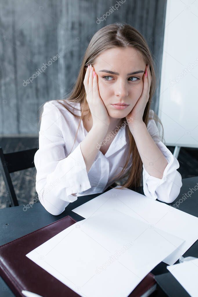Sad girl in a shirt sits at table in front of documents and wearily holds face in her hands. Monotonous and branch work in office.