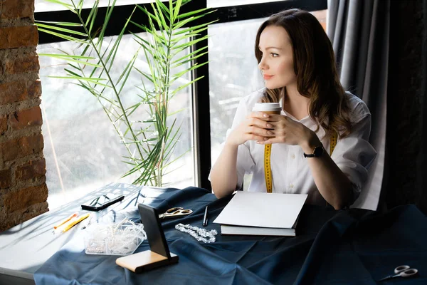 Fashion designer took a break and holds a coffee cup in her hands, sitting at a table with sewing materials and a smartphone.