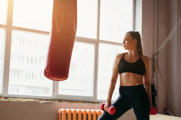 Vacker sportig tjej med röda hantlar i handen bredvid en boxningssäck i fitnessrummet med stora Windows — Stockfoto