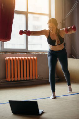 Spor kıyafetleri giyen bir atlet çevrimiçi pilates kursunda spor dambıllarıyla meşgul oluyor.