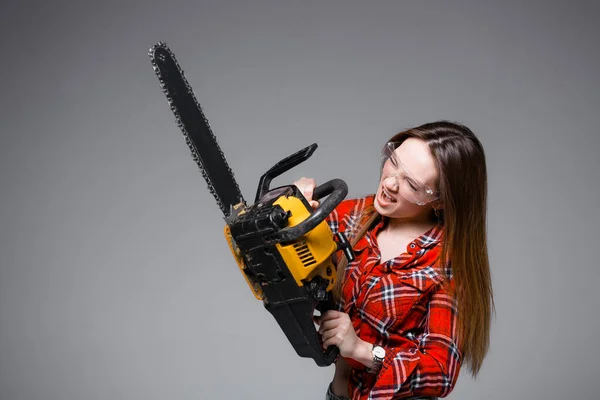 Beautiful sexy girl in red plaid shirt and in transparent glasses holds a chainsaw and crags. Advertising shot for hardware store. — Stock Photo, Image