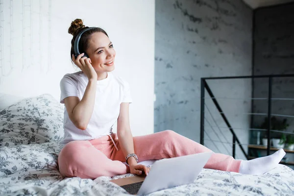 Гарненька руда усміхнена дівчина сидить на ліжку вдома з навушниками та слухає музику через ноутбук . — стокове фото