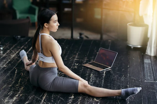 Fata este angajată să se întindă pe sfoară. Își întinde picioarele în lateral în timp ce stătea pe podea în fața laptopului. — Fotografie, imagine de stoc