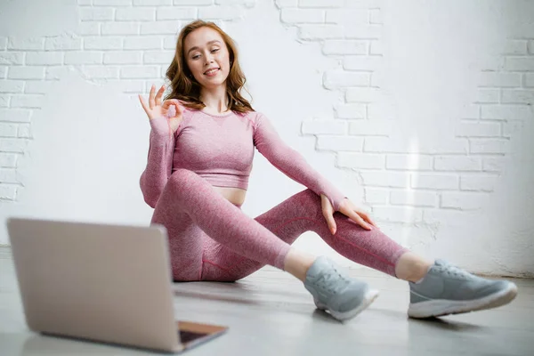 Trainer fitness într-un costum de sport roz face yoga pe o cameră web cu un student printr-un laptop și arată semnul bun cu degetele — Fotografie, imagine de stoc