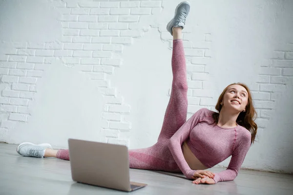 Yoga antrenor într-un costum roz predă yoga online pe un laptop și arată exerciții de ridicare a piciorului în timp ce se află pe podea — Fotografie, imagine de stoc