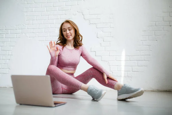 Рудоволоса дівчина в рожевому спортивному костюмі робить йогу на веб-камеру з тренером через ноутбук і показує правильний знак пальцями — стокове фото