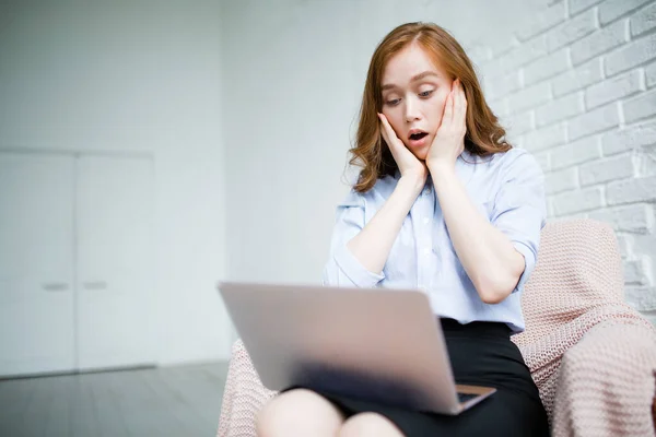 Chica de negocios mira con sorpresa en la pantalla del ordenador portátil y está sosteniendo con dos manos su cara de shock mientras está sentado en un sillón — Foto de Stock