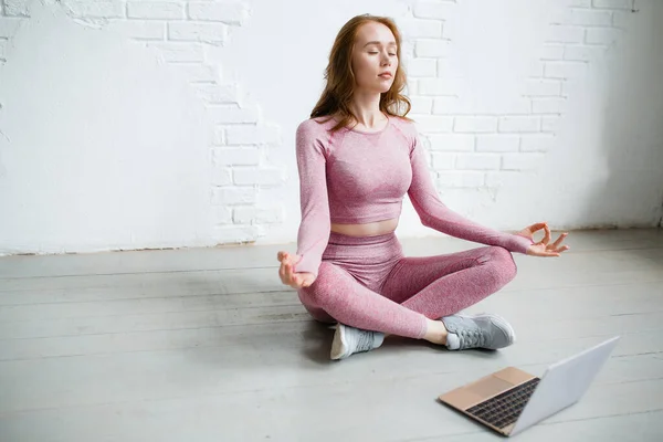 Fata cu părul roșu în tracksuit roz stă în poziția de lotus muzică relaxantă și video pe laptop în studio — Fotografie, imagine de stoc