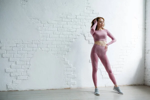 Entrenador niña pelirroja en un traje deportivo rosa se encuentra en pleno crecimiento sobre un fondo de una pared de ladrillo blanco en el estudio —  Fotos de Stock