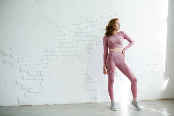 Chica pelirroja modelo entrenador en un traje deportivo rosa se encuentra en pleno crecimiento sobre un fondo de una pared de ladrillo blanco en el estudio —  Fotos de Stock