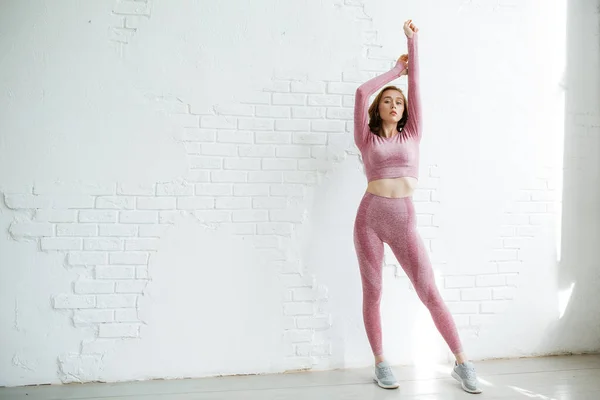 Chica-entrenador pelirroja en un chándal rosa se encuentra en pleno crecimiento estirando sus brazos contra una pared de ladrillo blanco en el estudio —  Fotos de Stock