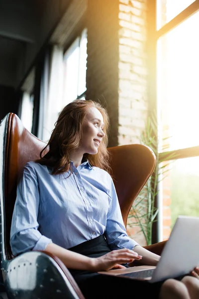 Červenovlasá dívka sedí v koženém křesle s notebookem na klíně a usmívá se na pozadí panoramatického okna. — Stock fotografie