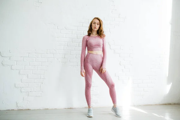 Entrenador niña pelirroja en un traje deportivo rosa se encuentra en pleno crecimiento sobre un fondo de una pared de ladrillo blanco en el estudio —  Fotos de Stock