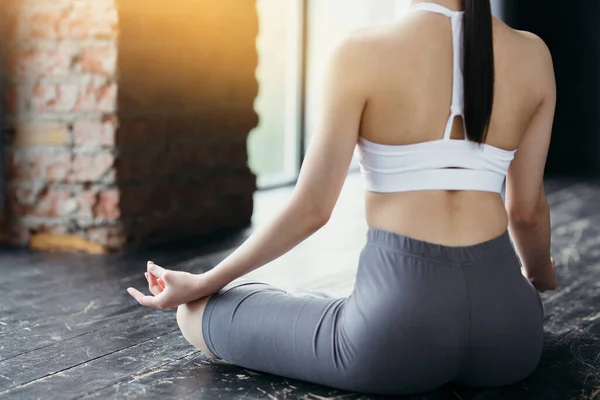 Vrouwelijke figuur achteraanzicht. Fitness trainer in een witte top en grijze legging zit in een lotuspositie in de zon op de vloer — Stockfoto