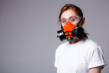 Beyaz tişörtlü güzel kızıl kız maske solunum cihazı ve güvenlik gözlüğü takıyor ve gri arka plandaki kameraya bakıyor.