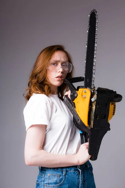 Close-up. Redhead woman in safety glasses in a white T-shirt and jeans holds a chainsaw near her face and looks at the camera. — Stock Photo, Image
