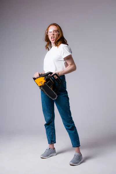 Chica pelirroja con una camiseta blanca y jeans sonríe amenazadoramente a la cámara y sostiene una motosierra sobre un fondo gris —  Fotos de Stock