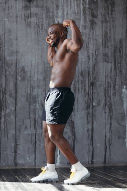 Çıplak gövdeli Afro-Amerikalı genç kaslı adam pazılarını gösteriyor. Gri bir arka planda güneşte duruyor