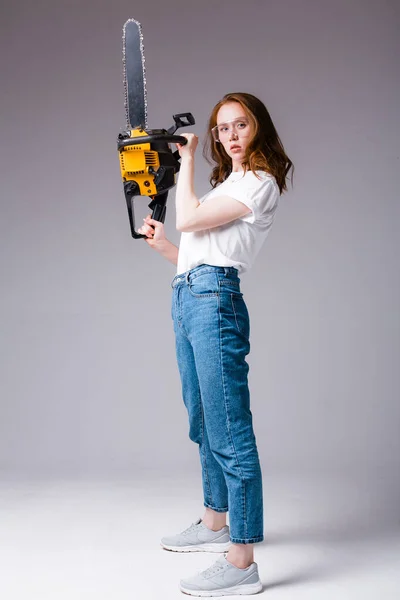 Chica pelirroja atractiva en una camiseta blanca y jeans sostiene una motosierra verticalmente y mira a la cámara sobre un fondo gris —  Fotos de Stock