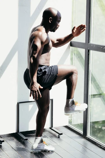 Koşudan önce kaslı erkek fitness antrenörü ısınma egzersizi yapar. Parlak bir stüdyoda büyük bir pencerenin yanında duruyor. — Stok fotoğraf