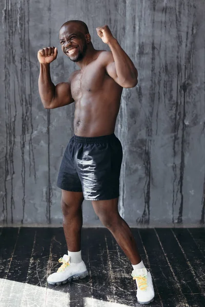 Hombre africano con una prensa llena de baches y un cuerpo musculoso en un estudio brillante, levanta las manos y se regocija — Foto de Stock