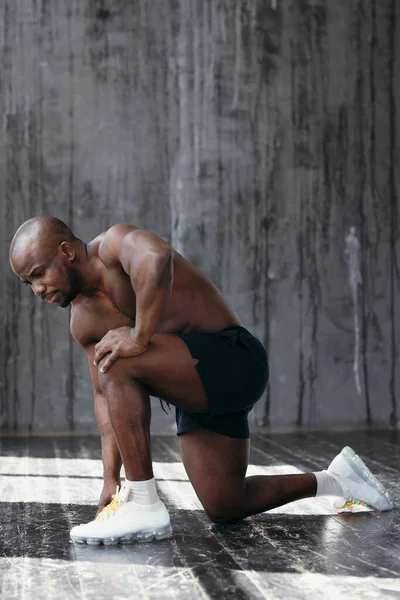 Un atleta negro se para sobre una rodilla, demuestra músculos. Desnudo afroamericano en pantalones cortos negros —  Fotos de Stock