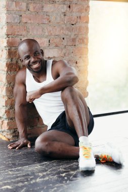Beyaz tişörtlü, siyah şortlu siyah kaslı bir adam spor salonunda yerde oturuyor ve gülümsüyor.