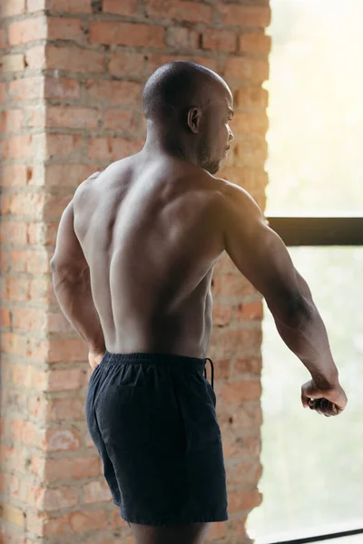 Muscular African American demonstrates back muscles while standing in front of a window.
