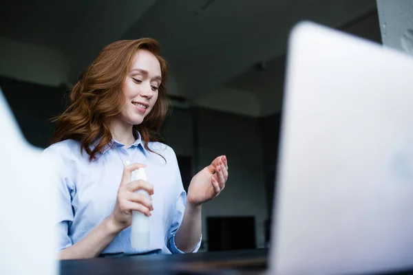 Američanka v modré košili si na ruce nasype dezinfekci. V popředí je kryt notebooku. Práce z domova. — Stock fotografie