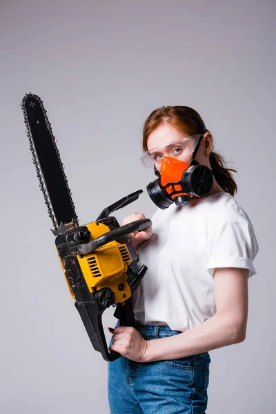 Chica pelirroja en camiseta blanca en respirador de construcción y gafas sostiene una motosierra. Banner publicitario para una ferretería —  Fotos de Stock