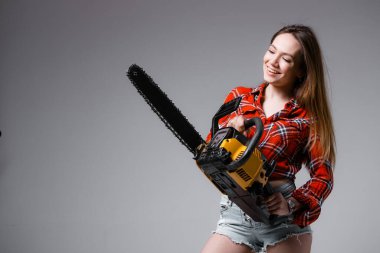 Kırmızı gömlekli seksi bir kız, elinde testere olan kot şortlu. Gri arka planda kadın inşaatçı